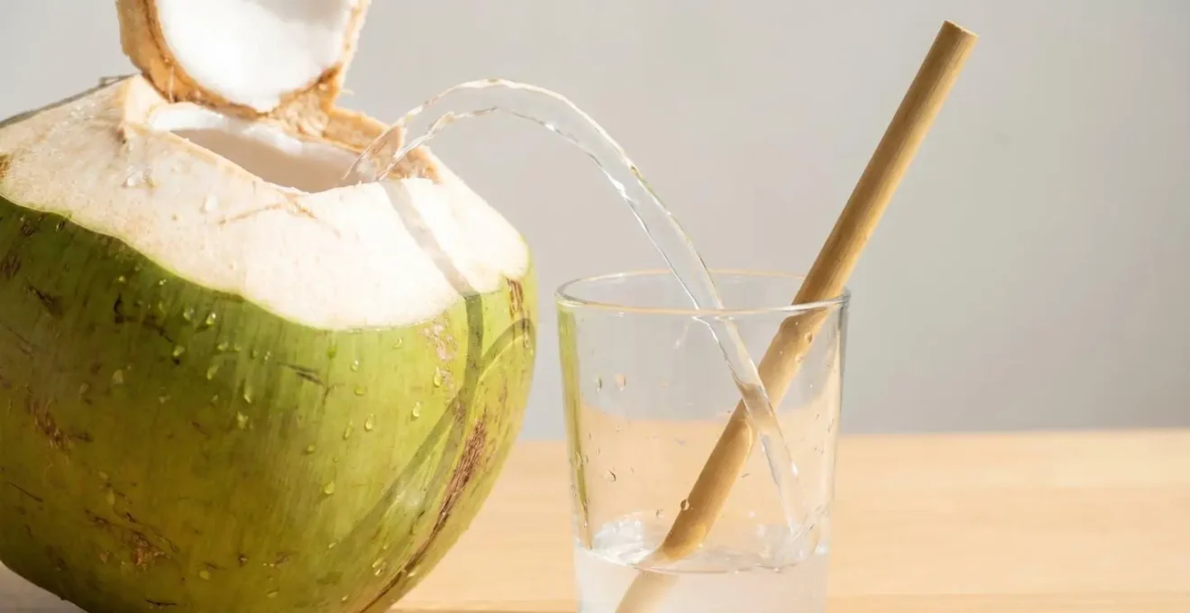 Une noix de coco fraîche coupée en deux avec de l'eau de coco transparente qui coule dans un verre, une paille en bambou naturel visible sur fond clair