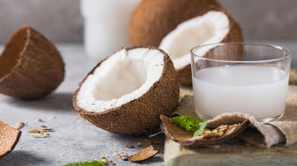 Une noix de coco fraîche coupée en deux avec de l'eau de coco transparente qui coule dans un verre, une paille en bambou naturel visible sur fond clair